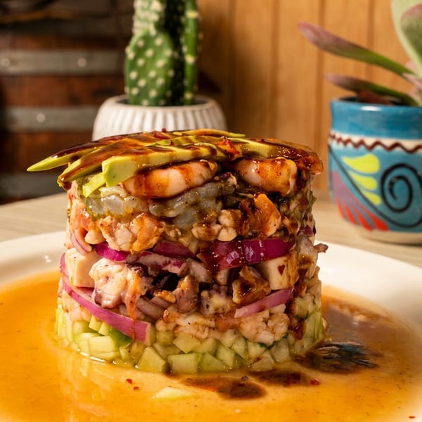 Stacked seafood ceviche with avocado, shrimp, red onion, and cucumber, drizzled with sauce.