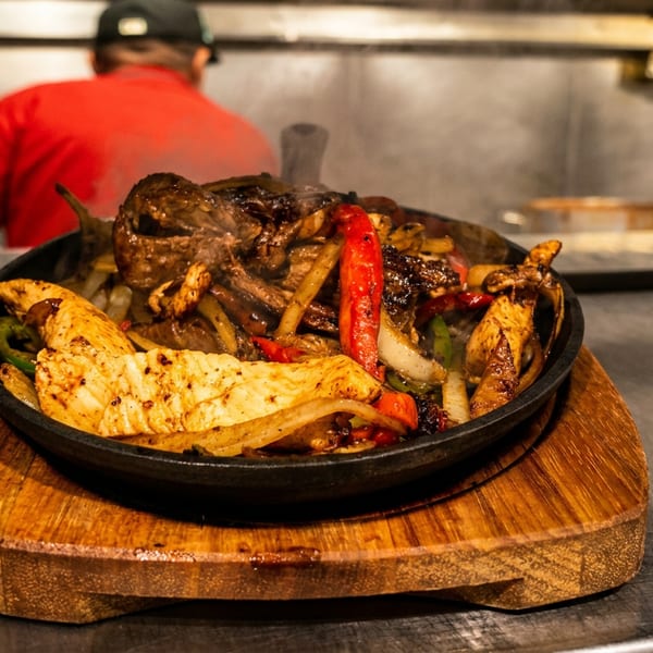 Sizzling fajitas with chicken, steak, peppers, and onions on a wooden board in a restaurant kitchen.