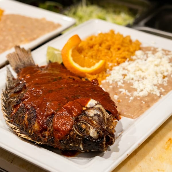 Whole fried fish covered in red sauce, served with rice, refried beans, and an orange slice.