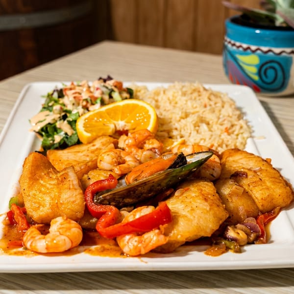 Seafood dish with fish, shrimp, mussels, rice, salad, and an orange slice on a white plate.