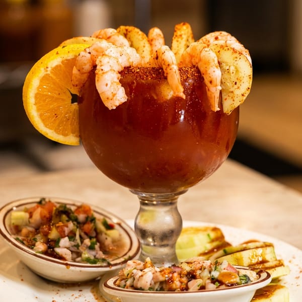 A large goblet of red cocktail with shrimp, cucumber, and orange slices, served with two small ceviche bowls.