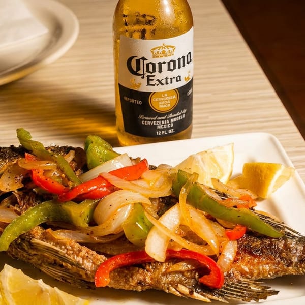 Fried whole fish topped with sautéed onions and bell peppers, with lemon wedges and a Corona beer.